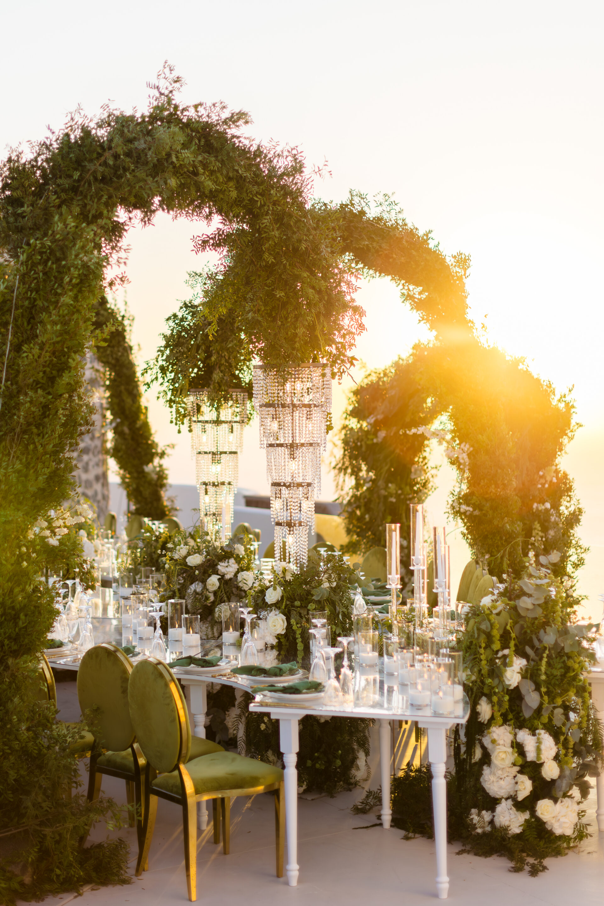 Elegant wedding reception table with crystal chandeliers and lush floral arches at sunset in Santorini