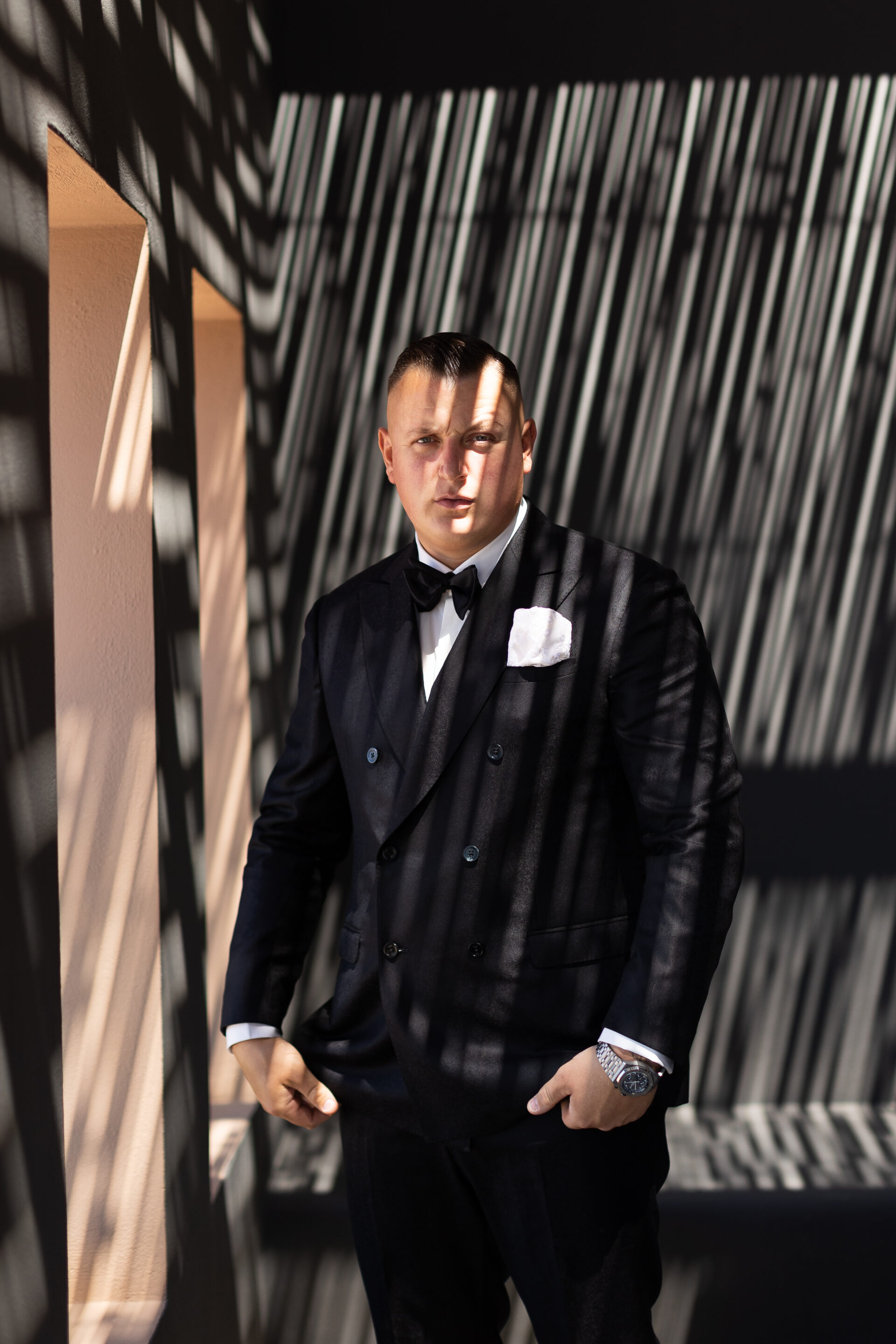 Groom in black tuxedo posing in dramatic striped shadow light in Santorini, captured by Santorini photographer