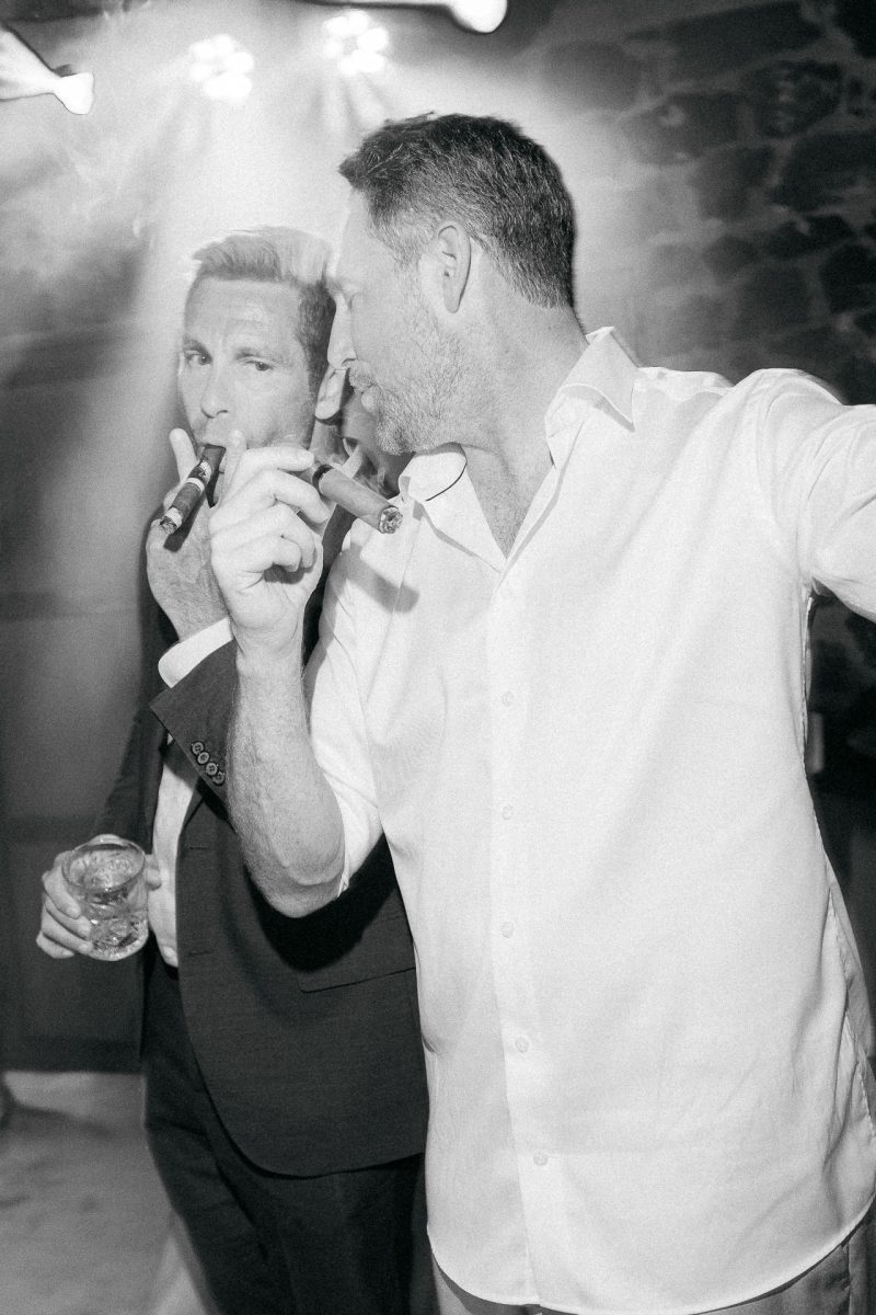 Santorini wedding photographer captures groom smoking a cigar with guest.