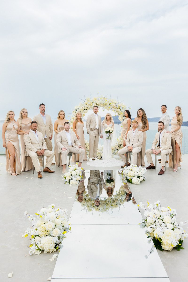Santorini wedding photographer captures full bridal party in front of floral arch at cliffside ceremony.